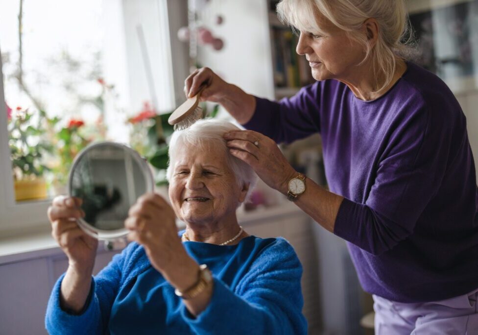 Woman brushing elderly woman's hair, holding mirror.